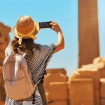 Traveler, traveling with Discovery Tours Egypt, taking a photo of ancient ruins in Egypt with a smartphone, wearing a straw hat, capturing Egypt’s rich history.